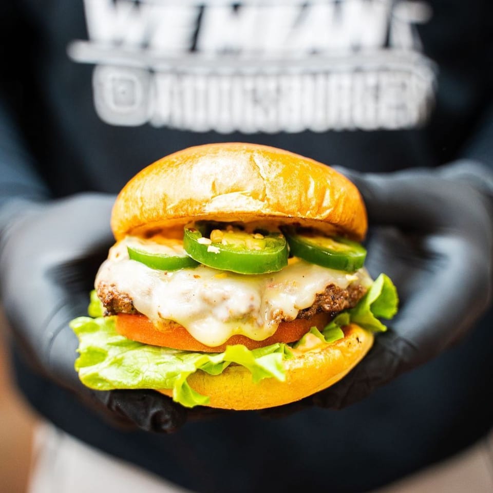 Gloved hands holding a Tee's A1 Dogs burger with melted cheese, jalapeños, lettuce, and tomato.