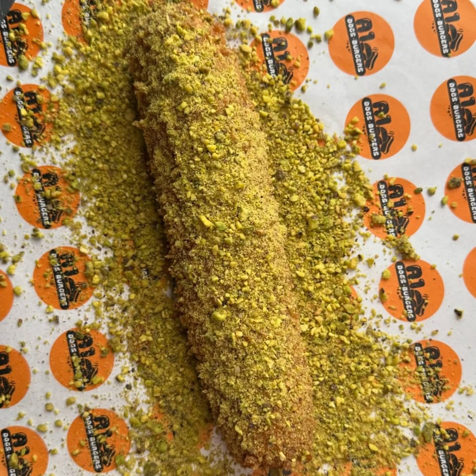 A Tee's A1 Dogs corn dog, heavily coated in bright green pistachio crumbs, on branded paper.