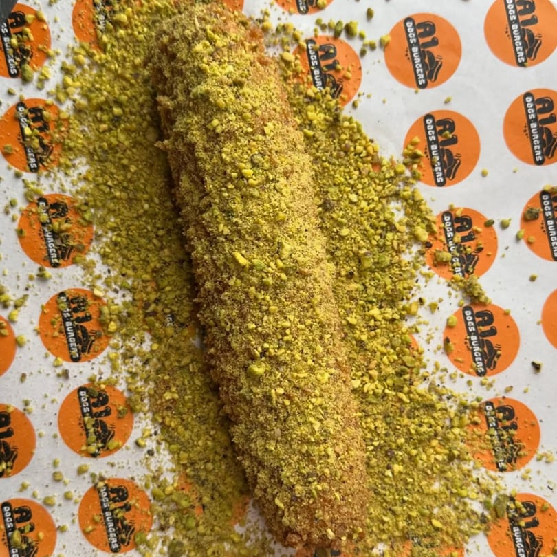 A Tee's A1 Dogs corn dog, heavily coated in bright green pistachio crumbs, on branded paper.
