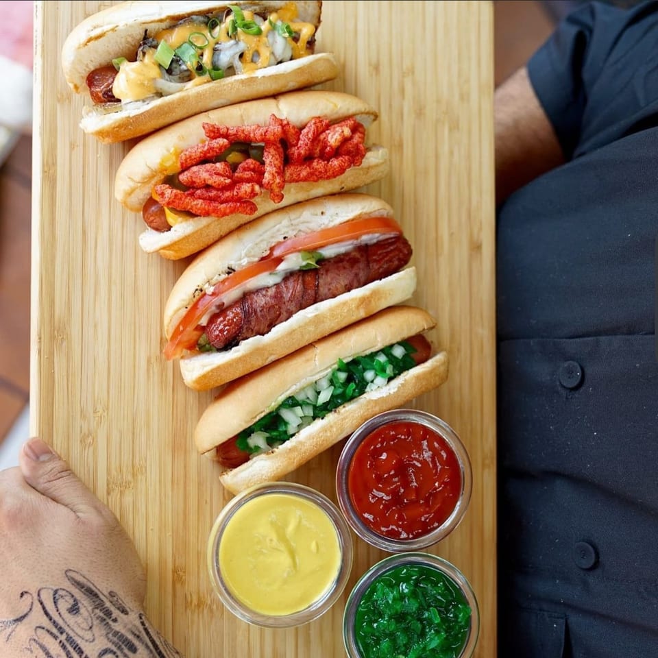 Five Tee's A1 Dogs hot dogs with various toppings and three dipping sauces on a wooden board.