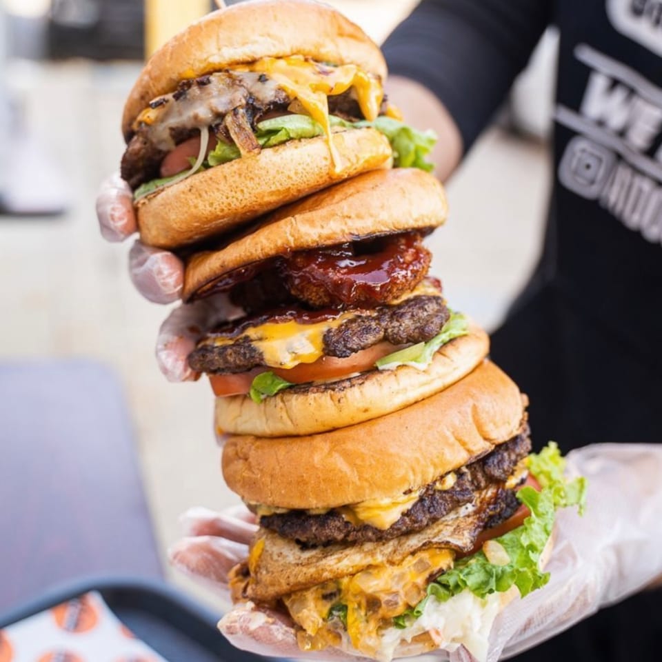 Gloved hands holding a towering stack of four Tee's A1 Dogs burgers with various toppings.
