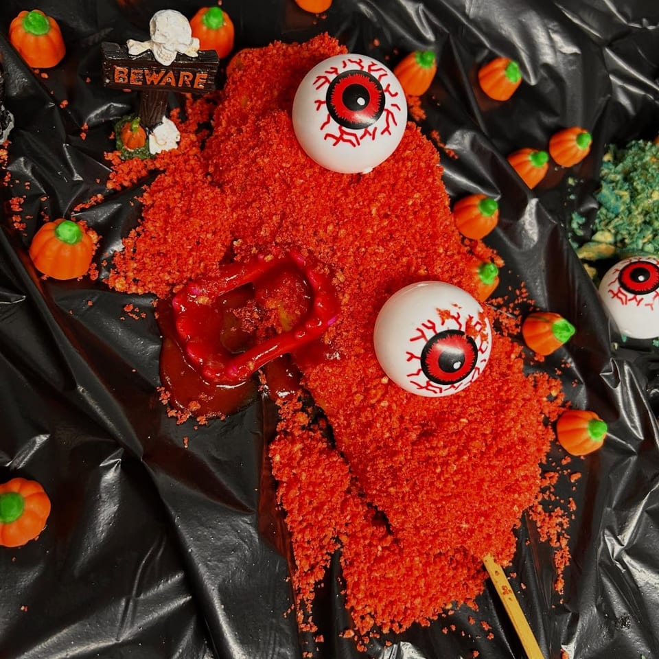 Halloween-themed hot dog with red crumbs, fake eyeballs, and a "Beware" sign on a black surface.